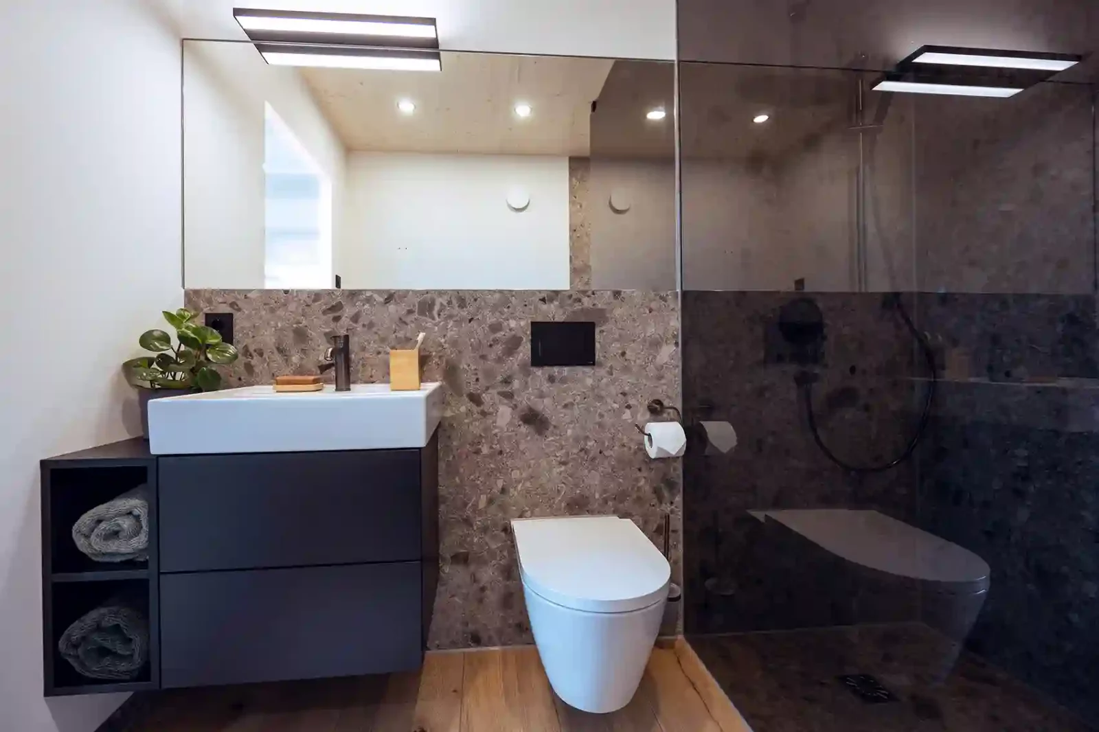 Modulbad Zermatt mit holzoptik Fliese und schwarzem Möbel einem aufgesetzten Waschtisch und offenen Regal mit Handtüchern.